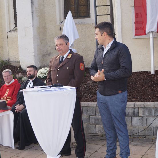 Zwei Männer stehen vor einem Tisch, einer in Uniform und der andere in einer Jacke. Sie scheinen zu sprechen. Hinter ihnen sitzen drei Männer auf einer Bank, einer trägt einen roten Umhang. Ein Flagge hängt an der Wand hinter den sitzenden Männern.