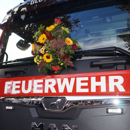 Ein Feuerwehrfahrzeug mit einem Blumenstrauß auf der Windschutzscheibe. Das Wort 'FEUERWEHR' steht in weiß auf einem roten Schild über dem Stoßfänger.