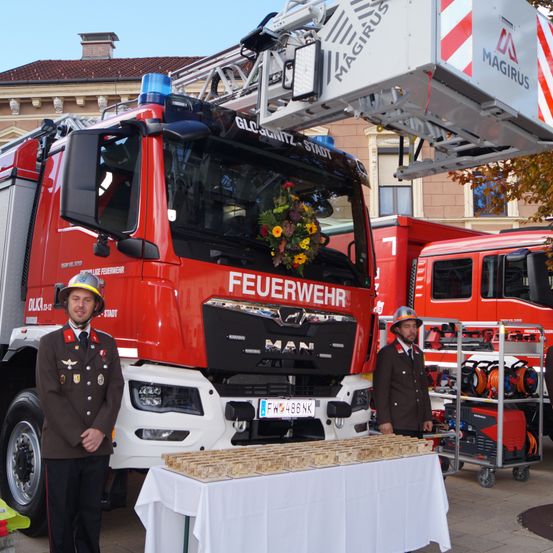 Zwei Feuerwehrleute stehen neben einem roten Feuerwehrauto mit einer Leiter darauf. Vor ihnen steht ein Tisch mit einigen Gegenständen.