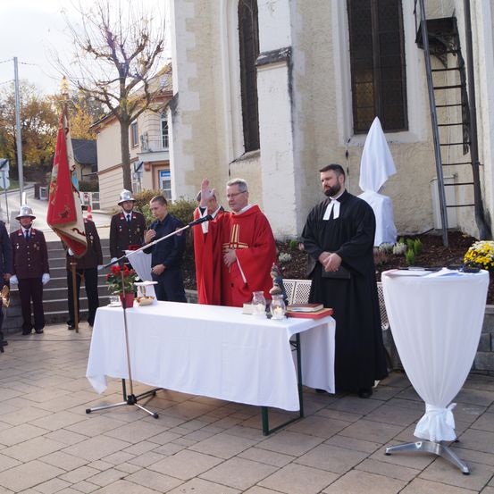 Ein Priester spricht in ein Mikrofon auf einem mit weißem Tuch bedeckten Tisch vor einer Kirche. Ein Mann in einem roten Gewand steht hinter dem Tisch.