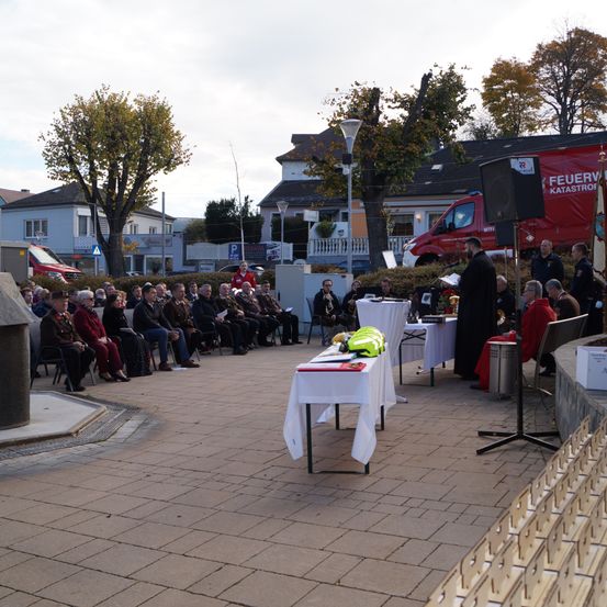 Eine Gruppe von Menschen sitzt auf Stühlen und Tischen um eine Bühne herum, mit einem Lautsprecher und einem roten Fahrzeug im Hintergrund. Eine Person steht und spricht.