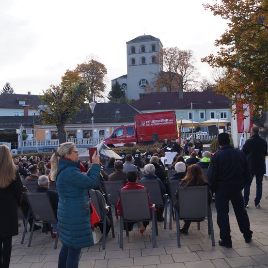 Eine Menschenmenge ist für eine Veranstaltung draußen versammelt. Eine Frau in einem blauen Mantel macht ein Foto. Ein rotes Fahrzeug ist im Hintergrund geparkt.