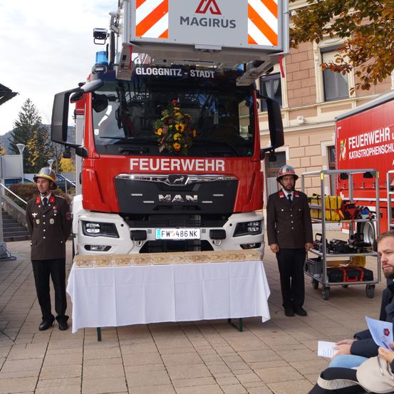 Zwei Feuerwehrleute stehen neben einem Feuerwehrauto mit einem Kranz auf der Front. Ein Tisch mit Auszeichnungen steht vor dem Auto. Ein Gebäude mit Fenstern und einem Baum ist im Hintergrund.