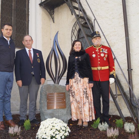 Vier Personen stehen vor einer Steinskulptur, möglicherweise bei einer Einweihungsveranstaltung. Ein Mann in einem roten Uniform steht neben einer Frau in einem Blumenrock.