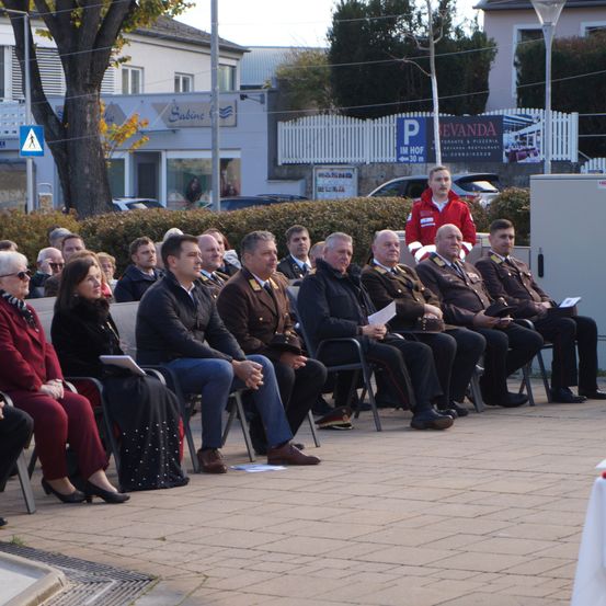 Eine Gruppe von Menschen, möglicherweise ein Komitee, sitzt in einer Reihe, einige halten Papiere. Ein Mann in einem roten Mantel steht vor ihnen und hält möglicherweise eine Rede. Hinter ihnen steht eine Reihe geparkter Autos, und ein Gebäude mit dem Schild 'Sabine' ist sichtbar.
