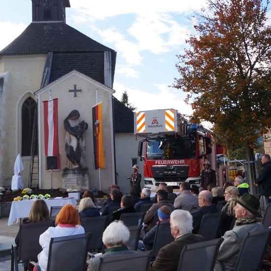 Ein Feuerwehrauto ist vor einer Kirche geparkt. Eine Statue steht vor der Kirche und zwei Fahnen befinden sich auf beiden Seiten der Statue. Eine Gruppe von Menschen sitzt auf Stühlen.