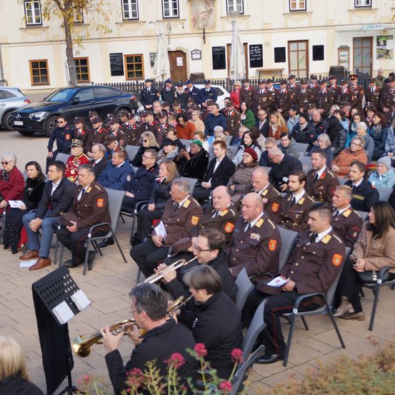 Eine große Menschenmenge sitzt in Stühlen im Freien vor einem Gebäude. Musiker spielen Trompeten, und eine große Gruppe uniformierter Personen steht hinter der sitzenden Menge.