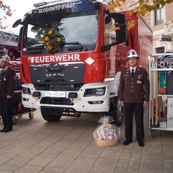 Ein Mann in Uniform steht neben einem roten Feuerwehrauto mit Blumen. Er steht vor einem Korb. Dahinter steht eine weitere Person in Uniform neben einem Wagen.