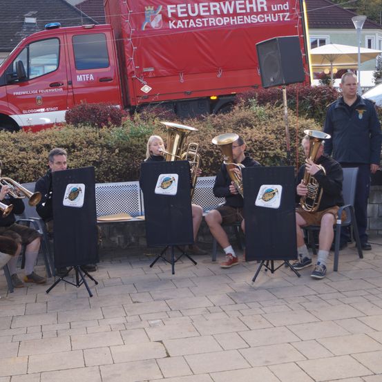 Eine Gruppe von Musikern spielt Trompeten vor einem roten Feuerwehrauto, mit Büschen und einem Lautsprecher im Hintergrund.