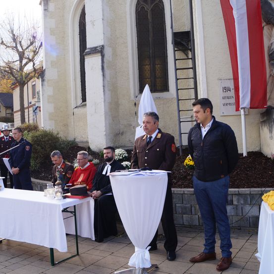Zwei Männer stehen vor einem Podium vor einer Kirche, mit Beamten und Geistlichen, die dahinter sitzen. In der Nähe sind eine Flagge und eine Statue zu sehen, und ein Gedenkstein ist an dem Gebäude angebracht.