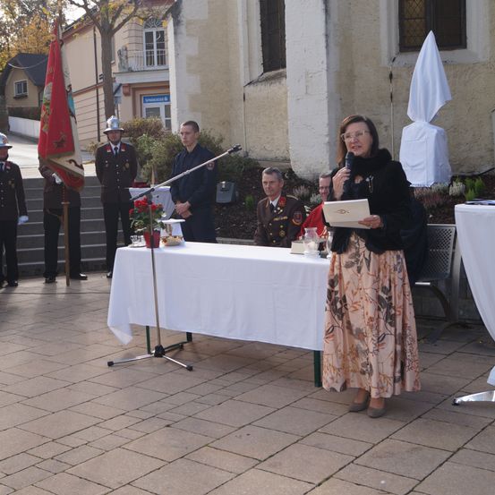 Eine Frau steht an einem Rednerpult, hält ein Buch in der Hand und spricht in ein Mikrofon. Hinter ihr steht ein Tisch mit weißer Tischdecke. Offiziere in Uniformen stehen um den Tisch herum. Ein Offizier hält eine Fahne.
