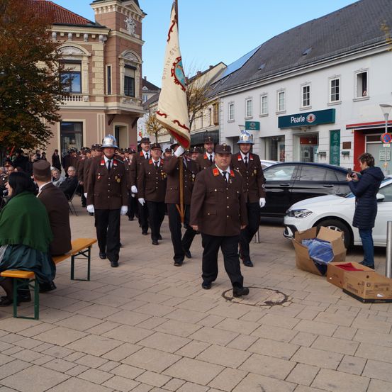 Eine Gruppe uniformierter Personen, möglicherweise Polizeibeamte, marschiert bei einer Parade mit einer Fahne. Zuschauer sitzen auf Bänken. Gebäude säumen die Straße, eines mit einem Schild mit der Aufschrift Pearle. Eine Person fotografiert die Szene vom Bürgersteig aus.