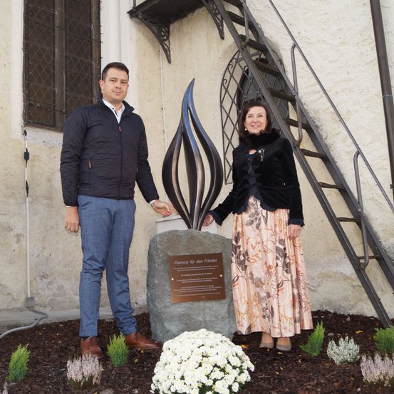 Ein Mann und eine Frau stehen lächelnd vor einem Denkmal mit einer Flammenskulptur, Blumen und einer Steintafel.