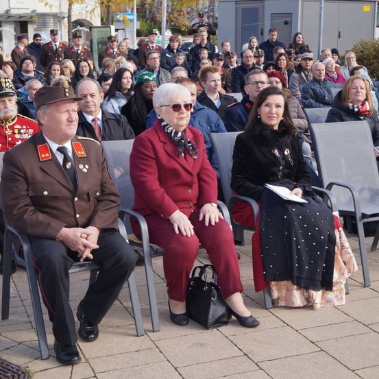Eine Menschenmenge sitzt in Stühlen, wahrscheinlich bei einer Veranstaltung. Ein Mann in Uniform und eine Frau in Rot sitzen in der vordersten Reihe.