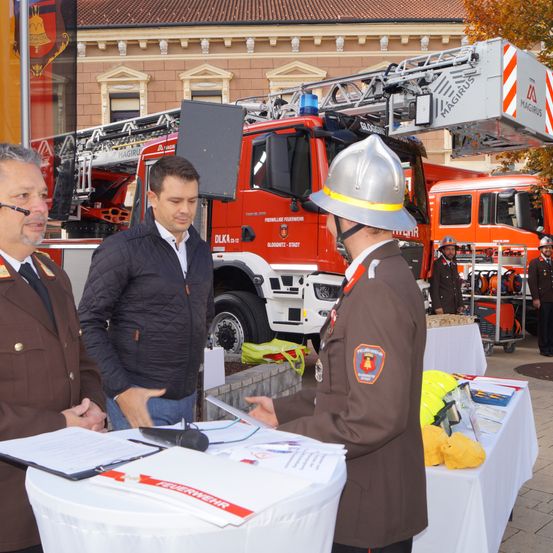 Drei Männer stehen in der Nähe eines Feuerwehrautos, wobei ein Mann in Uniform einem anderen Mann ein Papier überreicht. Ein Banner ist im Hintergrund.