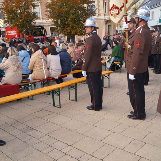 Militäroffiziere stehen neben hölzernen Bänken auf einem öffentlichen Platz, mit einer Menge, die auf den Bänken hinter ihnen sitzt. Bäume und Gebäude sind im Hintergrund.