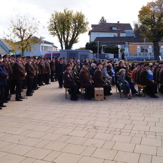 Eine Gruppe von Menschen in Militäruniformen steht in Formation. Mehrere Personen sitzen auf Stühlen vor ihnen. Ein Kasten steht auf dem Boden. Eine Straßenlaterne steht rechts. Einige Bäume stehen dahinter.