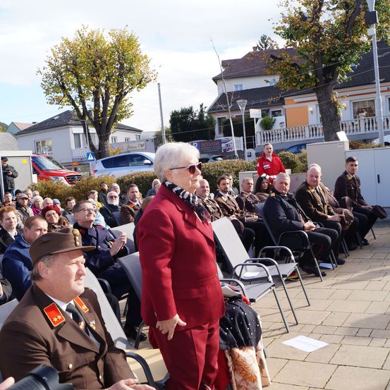 Eine Gruppe von Menschen in formeller Kleidung versammelt sich draußen in einer städtischen Umgebung, einige sitzend und andere stehend, mit Gebäuden und Autos im Hintergrund.