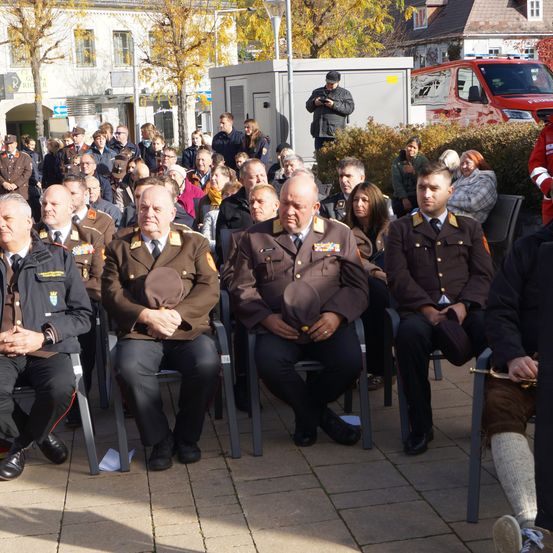Eine Gruppe uniformierter Personen sitzt in einer Reihe auf Stühlen, wahrscheinlich bei einer formellen Veranstaltung. Einige tragen Hüte, während andere sie halten. Ein Fotograf steht in der Nähe und hält die Szene fest.