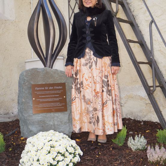 Eine Frau steht neben einem großen Stein mit einer Plakette, Blumen und einer Metallskulptur. Die Frau trägt eine schwarze Samtjacke und einen Blumenrock.