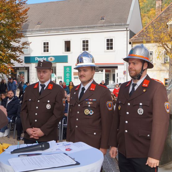 Drei Feuerwehrleute in Uniform stehen vor einem Tisch mit einem Mikrofon. Sie sind draußen in der Nähe einer Menschenmenge, mit Gebäuden und Bäumen im Hintergrund.