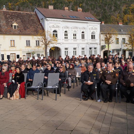 Eine Menschenmenge, möglicherweise bei einer militärischen Zeremonie, sitzt in Reihen von Stühlen im Freien vor einem weißen Gebäude.