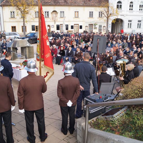 Eine Gruppe uniformierter Personen steht mit Fahnen vor einem Gebäude. Eine Menschenmenge ist versammelt, einige sitzend, andere stehend. Musikinstrumente sind aufgestellt, mit einem Lautsprecher in der Nähe.
