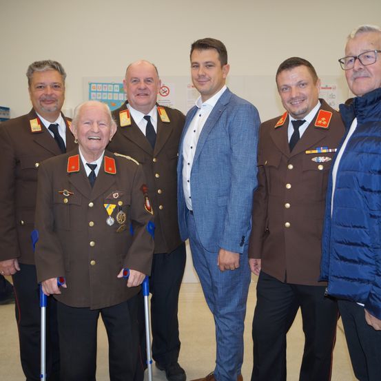 Fünf Männer in Militäruniformen und ein Mann in einer blauen Jacke stehen gemeinsam in einem Raum. Sie alle lächeln für die Kamera.