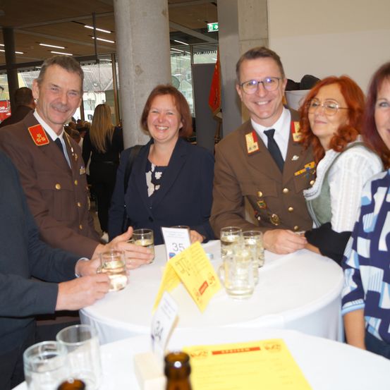 Eine Gruppe von Menschen, darunter zwei Männer in Militäruniformen, sitzen bei einer Veranstaltung um einen Tisch herum, lächeln und halten Gläser mit Wasser in den Händen.