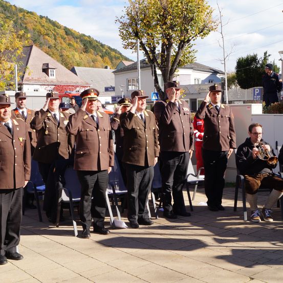 Eine Gruppe uniformierter Männer salutiert draußen. Ein Mann mit Brille spielt Trompete. Im Hintergrund sind Gebäude und Bäume zu sehen.