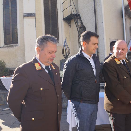 Drei Männer in Uniform stehen vor einem Gebäude. Der Mann in der Mitte trägt eine schwarze Jacke. Der Mann auf der linken Seite hat eine braune Uniform mit goldenen Knöpfen. Der Mann auf der rechten Seite trägt einen braunen Mantel mit Krawatte.