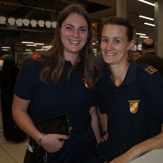 Zwei lächelnde Frauen in Uniform, eine hält ein Tablet, stehen nebeneinander. Sie sind bei einer Veranstaltung, mit einem verschwommenen Publikum und Kleidung im Hintergrund.