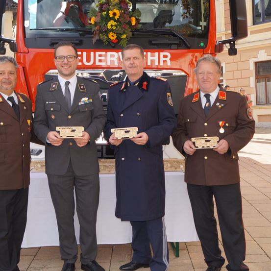 Vier Männer in Uniformen stehen vor einem Feuerwehrauto. Sie halten Holzplaketten in den Händen. Dahinter befindet sich ein Gebäude mit einem Fenster.