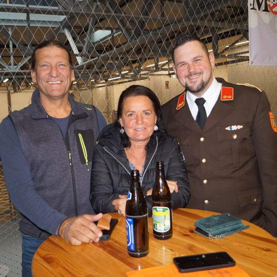 Drei Menschen, die in einem metallgeschützten Bereich lächeln und für ein Foto posieren, eine Frau mit zwei Bierflaschen vor sich und ein Mann in Uniform.