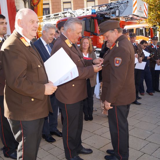 Ein Mann in Uniform überreicht einem anderen Mann in Uniform eine Auszeichnung, während andere hinter ihnen stehen. Im Hintergrund ist ein Feuerwehrwagen zu sehen.