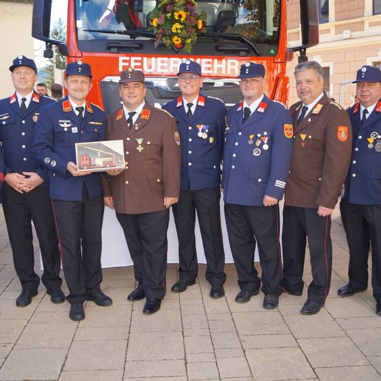 Eine Gruppe von Feuerwehrleuten in Uniform, geschmückt mit verschiedenen Medaillen, steht vor einem Feuerwehrwagen. Ein Mitglied hält ein gerahmtes Bild eines Feuerwehrwagens.