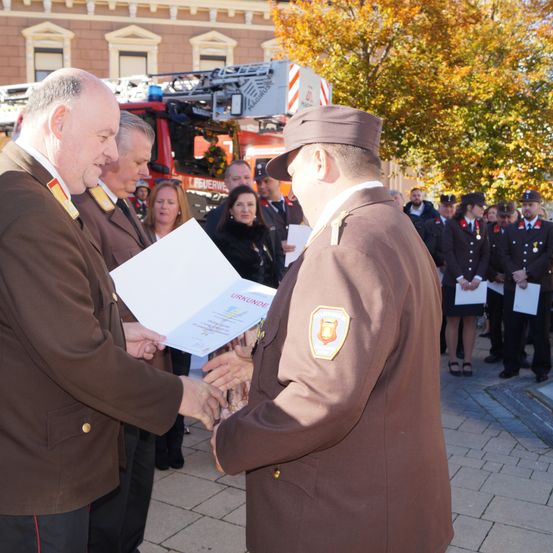 Zwei Männer in Uniformen tauschen bei einer Zeremonie im Freien ein Zertifikat aus, mit anderen hinter ihnen. Ein Feuerwehrauto ist im Hintergrund geparkt.
