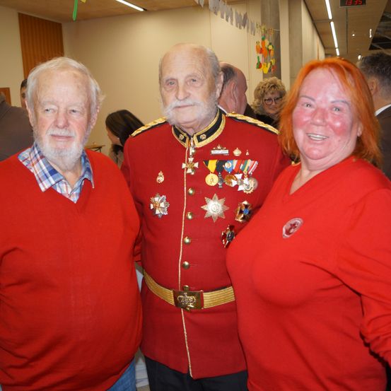 Zwei ältere Männer und eine Frau, alle in Rot, stehen zusammen und lächeln in die Kamera. Ein Mann hat Medaillen an seiner Uniform.