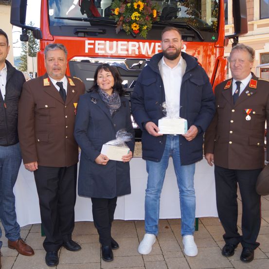 Fünf Personen stehen vor einem Feuerwehrauto. Ein Mann in einem weißen Hemd hält ein Geschenk und eine Frau in einem blauen Mantel hält ein verpacktes Geschenk. Dahinter bedeckt eine weiße Tischdecke einen Tisch.