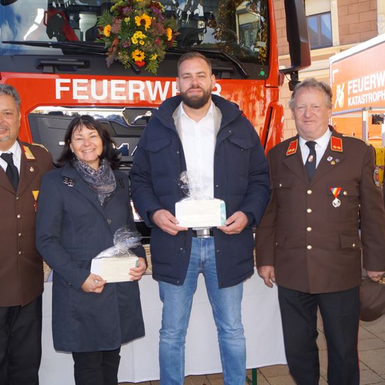 Drei Personen stehen vor einem Feuerwehrauto. Ein Mann hält ein Geschenk, während zwei andere lächeln, einer trägt eine Schürze.