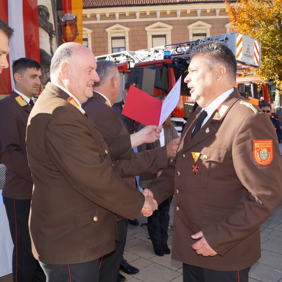 Zwei Männer in Militäruniformen schütteln sich die Hände, einer hält einen roten Ordner. Sie stehen vor einem Feuerwehrauto mit einem orange-weißen Farbschema.