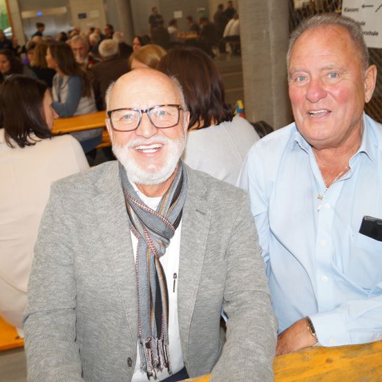 Zwei Männer sitzen an einem Tisch und lächeln für ein Foto. Der Mann auf der linken Seite trägt eine Brille und einen Schal. Der Mann auf der rechten Seite trägt ein hellblaues Hemd. Hinter ihnen sitzen weitere Personen an Tischen.