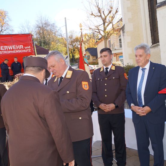 Eine Gruppe in Uniform steht vor einem Gebäude mit einem Banner. Ein Mann in Uniform befestigt eine Auszeichnung auf der Jacke eines anderen.