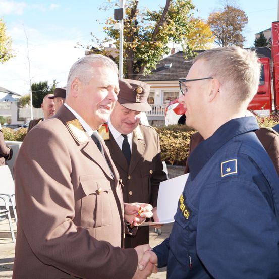Ein formeller Händedruck zwischen zwei Männern, einer in Uniform und der andere in einem dunkelblauen Anzug. Hinter ihnen hält ein Mann ein Stück Papier und einen Stift.