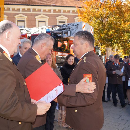 Drei Männer in braunen Uniformen, einer mit einer roten Mappe, unterhalten sich miteinander. Hinter ihnen steht eine Menschenmenge, darunter eine Frau mit einem Blumenkleid, in der Nähe eines Feuerwehrwagens und eines Gebäudes.