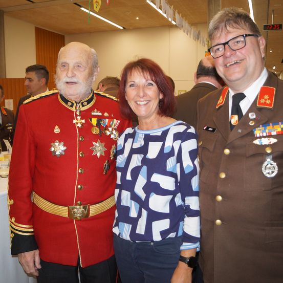 Drei Menschen stehen lächelnd für ein Foto zusammen. Der Mann auf der linken Seite trägt einen roten Militäruniform mit Medaillen, und die Frau in der Mitte trägt ein blaues Shirt. Der Mann auf der rechten Seite trägt eine braune Militäruniform.
