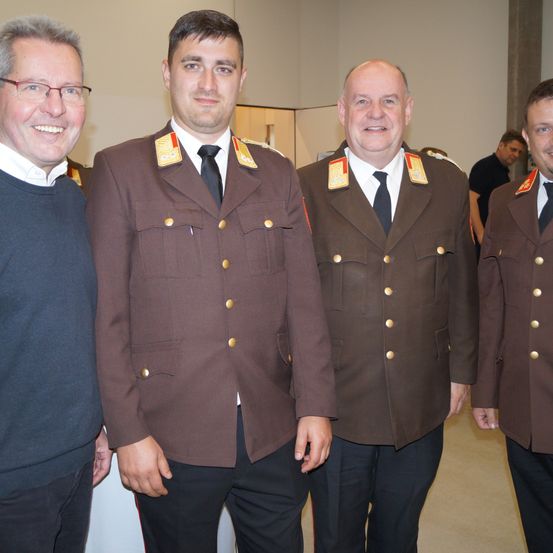 Vier Männer in Militäruniformen stehen zusammen und lächeln für ein Foto. Hinter ihnen sind zwei weitere Männer zu sehen, von denen einer ein schwarzes Shirt trägt.