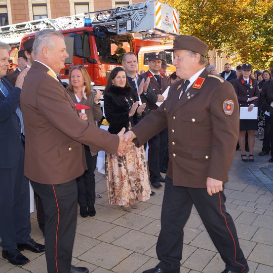 Zwei Männer in braunen Uniformen schütteln sich die Hände, umgeben von Zuschauern. Hinter ihnen steht ein Feuerwehrauto geparkt. Einige Leute klatschen.