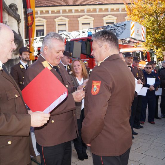 Eine offizielle Veranstaltung mit Feuerwehrleuten und Beamten in braunen Uniformen. Ein Mann hält eine rote Mappe, die er wahrscheinlich einem anderen Mann überreicht. Hinter ihnen halten Menschen Papiere. Ein Feuerwehrwagen ist sichtbar.