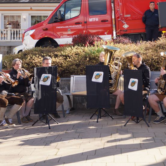 Eine Gruppe von Menschen spielt Musikinstrumente im Freien, mit einem Feuerwehrauto im Hintergrund.